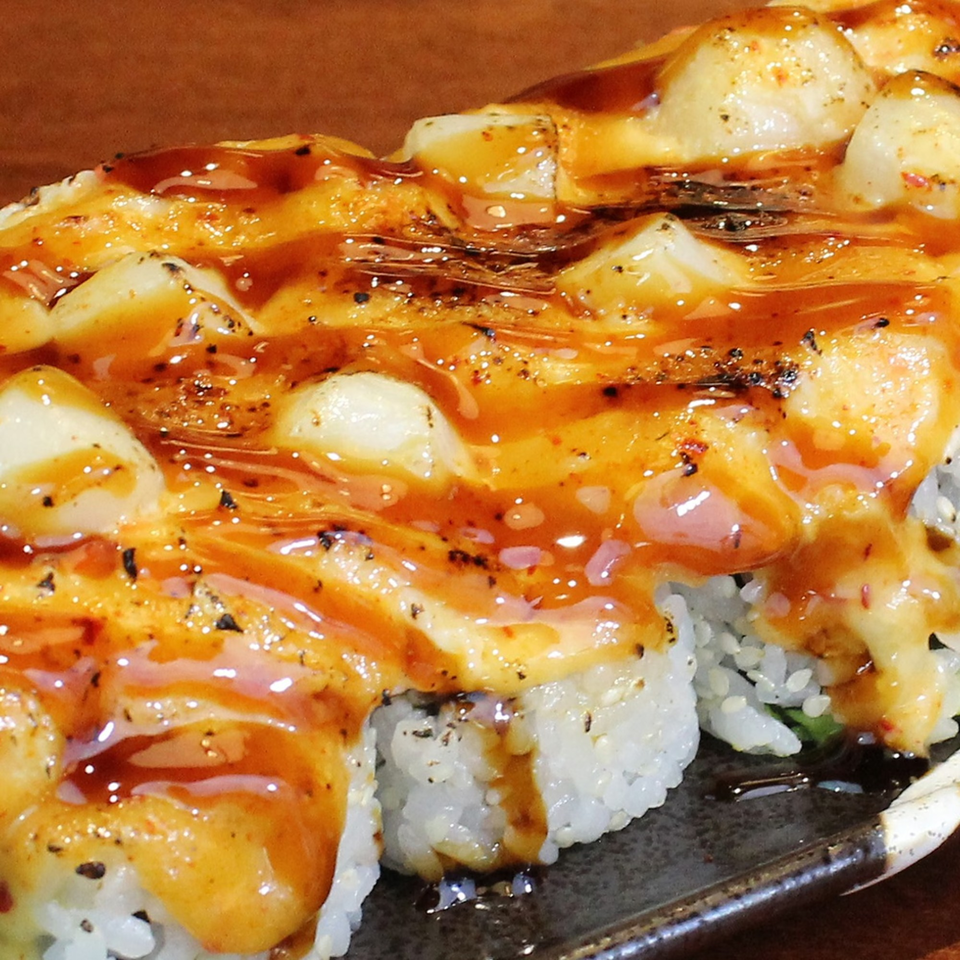 Close-up of a sushi roll topped with baked scallops and a rich, glossy sauce.