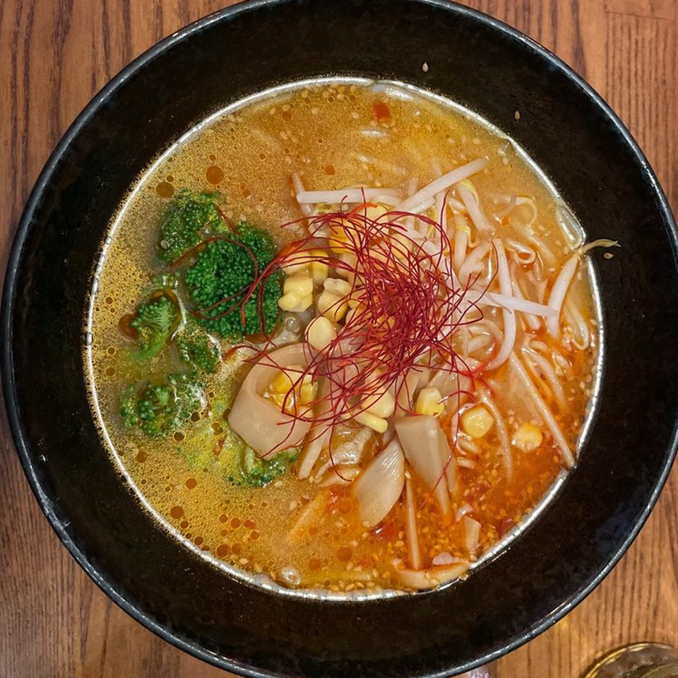 Overhead view of a ramen bowl with broth, broccoli, corn, bean sprouts, and red chili threads.